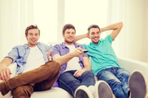three men sit on a couch with their feet one, one raises a TV remote